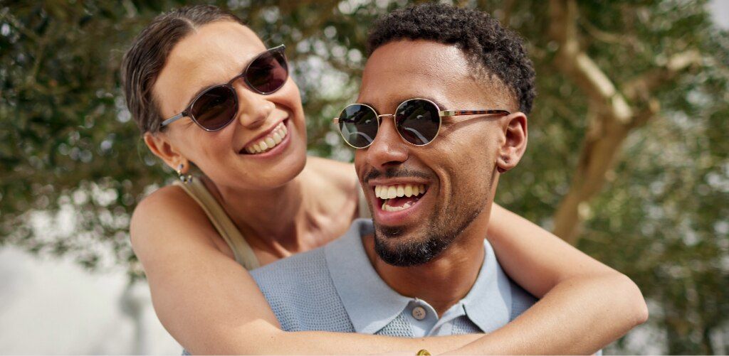A smiling couple enjoys a sunny moment outdoors, both wearing sunglasses. The woman has brown hair tied back, wearing small oval frames, leans over the man’s shoulders with her arms around him, while he wears round metal sunglasses with tortoiseshell acet