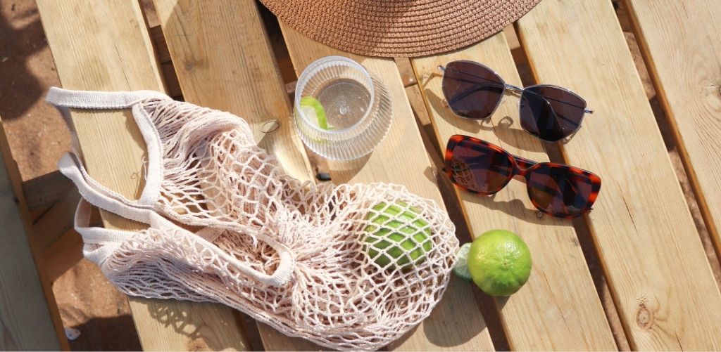 A sunny outdoor scene shows a wooden table with summer accessories neatly arranged. There’s a woven straw hat, two pairs of sunglasses (one with silver geometric lenses and another with bold tortoiseshell frames), and a mesh tote bag holding green limes. 