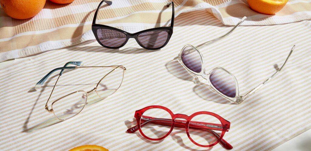 A selection of sunglasses and eyeglasses arranged on a striped cloth in bright sunlight. The styles include black cat-eye shades, gold metal cat-eye glasses, clear-framed square sunglasses, and bold red round frames. Oranges are placed around the scene, a