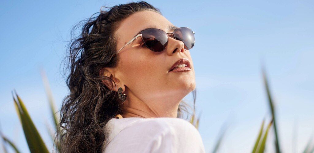 A woman with wavy brown hair wears slim, oval sunglasses with tinted lenses, tilting her face upward toward the sunlight. She has on a white top and decorative earrings, with a clear blue sky and hints of greenery in the background, creating a bright, sum