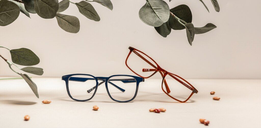 Two pairs of Glasses Direct bio-nylon eyeglass frames are displayed on a neutral beige surface beneath decorative eucalyptus leaves. The Louis blue square frame sits in the foreground, while the Leslie translucent amber-brown rectangular frame is propped 