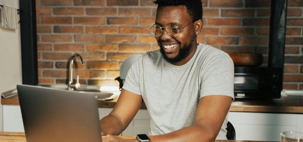 A young man with short black hair wearing round metal glasses smiling brightly. He is sat down in front of a brick wall and kitchen counter with a sink and microwave in the background. The man is looking at the laptop before him.