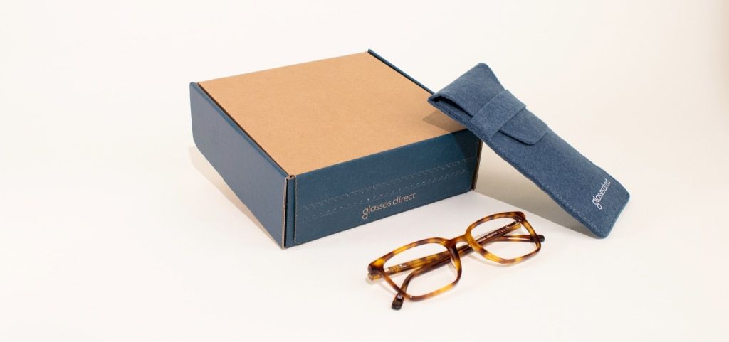 A cardboard box with a soft glasses case resting upwards on it. A pair of tortoiseshell acetate glasses are folded up in front.