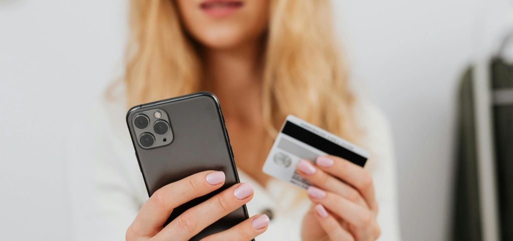A close-up of a ladies hands holding a mobile phone before her and a credit card in the other hand. The bottom of her face and shoulders are blurred in the background.
