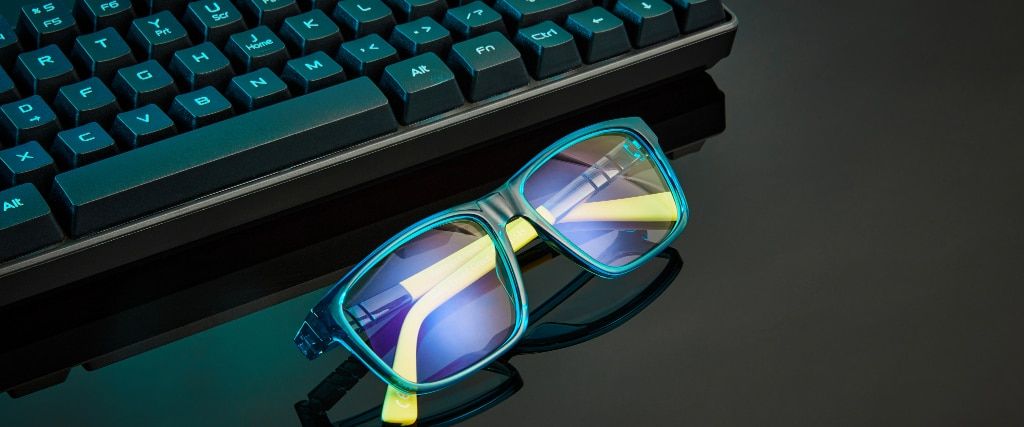 Close up of a pair of blue and yellow square glasses folded on a black shiny surface near a keyboard.