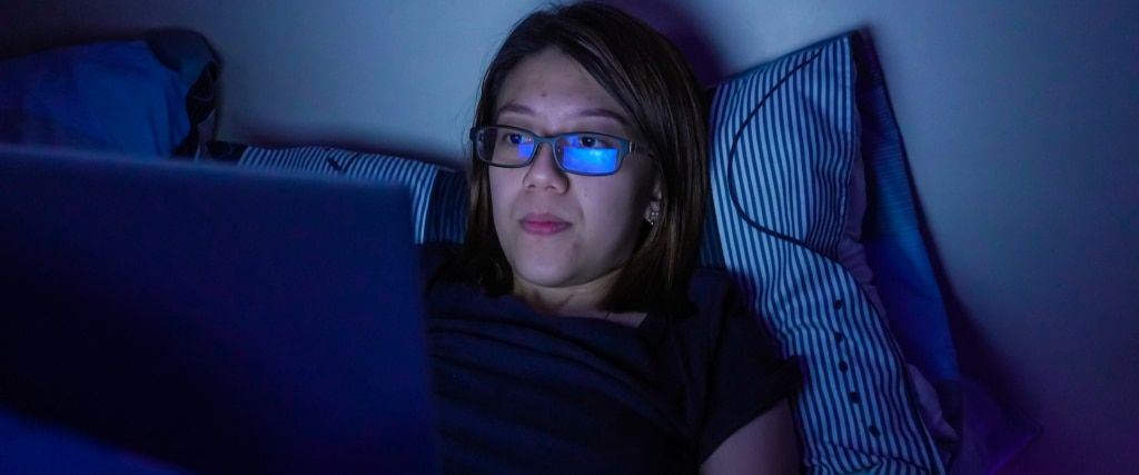 A young woman with a brown bob sits in bed in a dimly lit room. She wears a dark blue t-shirt and rectangular acetate glasses with blue light lenses, gazing at the laptop screen in front of her.