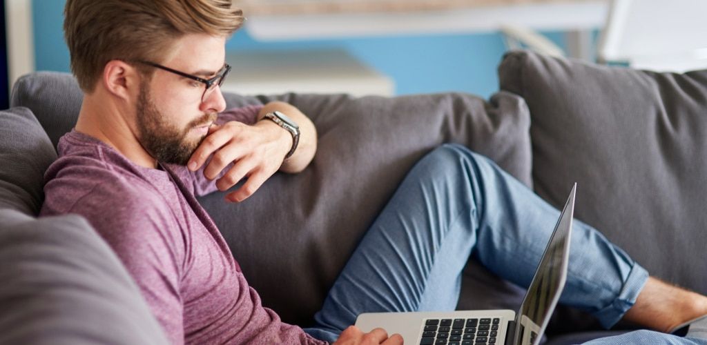 A man wearing rectangular glasses relaxes on a grey sofa while using a laptop. Dressed in a casual burgundy t-shirt and light blue trousers, he rests one hand thoughtfully against his chin as he focuses on the screen. The softly lit home setting and comfo