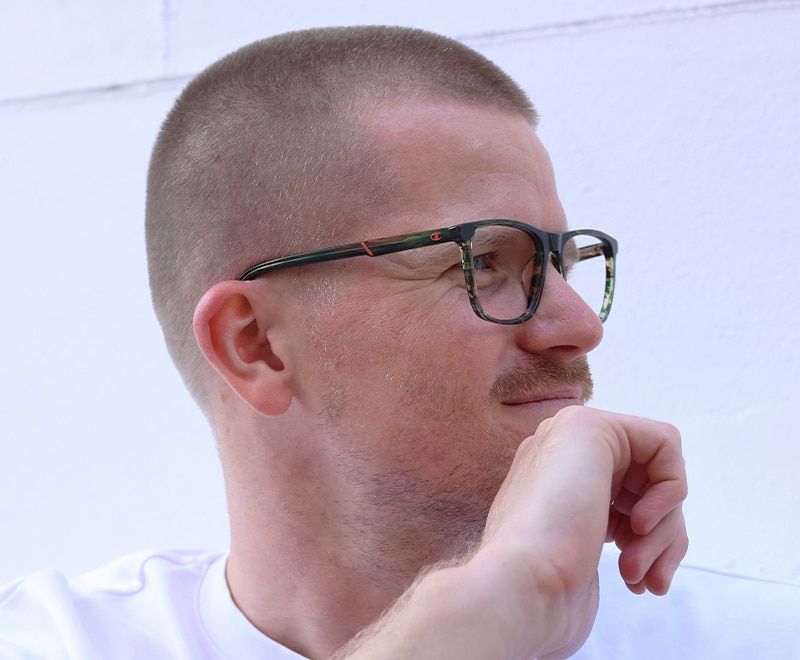 Young man wearing green tortoiseshell, round Champion frames outdoors looking to the right