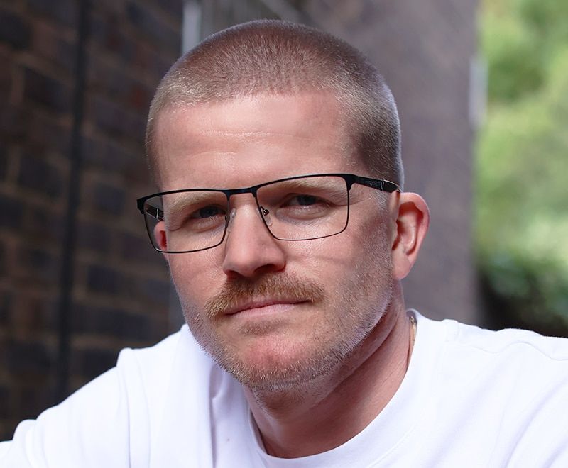 Young man wearing grey metal, rectangular Champion frames outdoors looking directly at camera