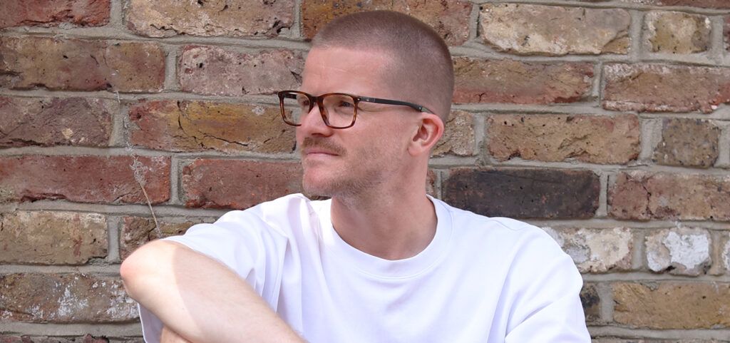 A portrait shot of a young man with a short buzz cut and light facial hair, sitting with one arm resting casually on his knee, in front of a brick wall of mixed beige and red. He is wearing a plain white tee and a pair of square acetate sports glasses whi