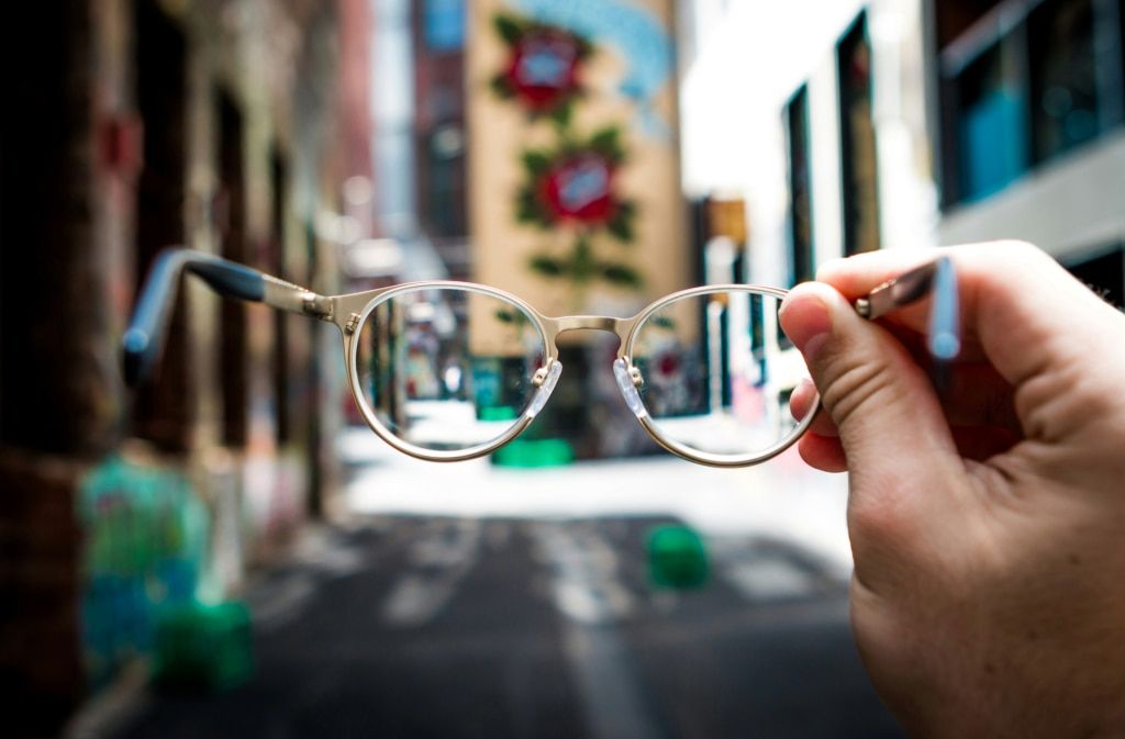 A close up of a man's hand holding a pair of gold metal glasses. The background is blurred and appears to be a cityscape.