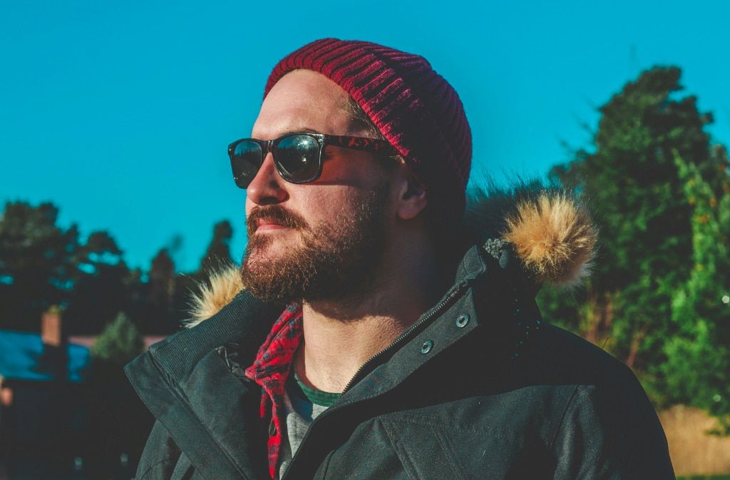 A young bearded man wearing a red beanie, acetate sunglasses and a parka coat looking in the distance. He is standing in an outdoor setting. Trees and a house with a chimney can be seen in the background and the sky is bright blue.