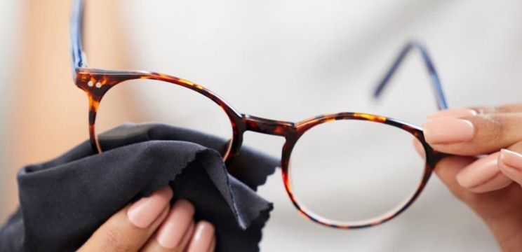 A close up of a hand cleaning a pair of tortoiseshell glasses with a black glasses cloth.