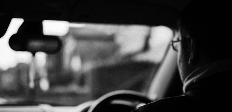 A black and white image showing the back of a man from the left-hand side. He is wearing glasses and a coat and is driving a car. The rear-view mirror, windscreen and steering wheel can be seen blurred before him.
