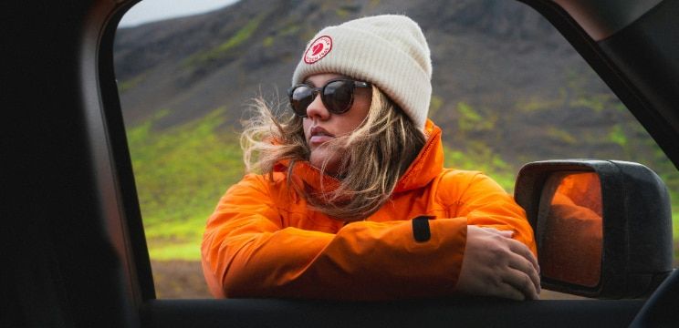 A young blonde woman wearing a cream-coloured beanie, black sunglasses and a bright orange coat. She is leaning on the open window of a car and appears to be in a mountainous outdoor setting.