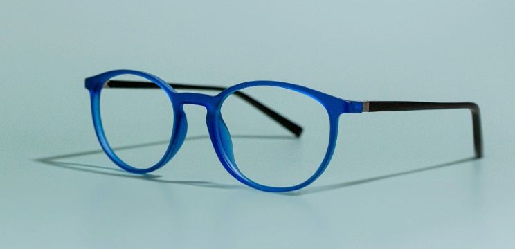 A round acetate frame which is blue at the front with black temples. It is propped open to one side on a bright blue background.