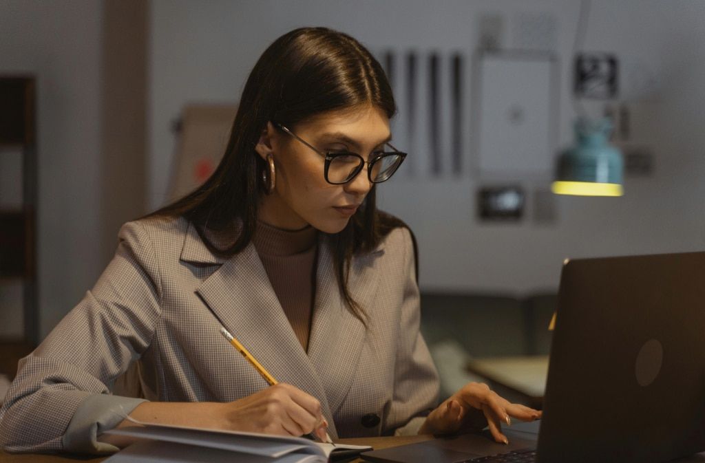 A young lady with long, straight dark hair moving the mouse pad on a laptop and writing notes in a notebook. She is in a bright, modern indoor setting and is wearing a checked blazer, brown polo neck, golden hoop earrings and black acetate glasses with si