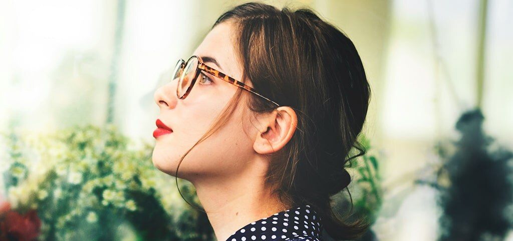 A side shot of a young lady with brown hair tied up in a loose bun. She appears to be in an indoor setting with flowers out of focus in the background. She is looking dreamily into the distance. The lady is wearing bright red lipstick, tortoiseshell glass