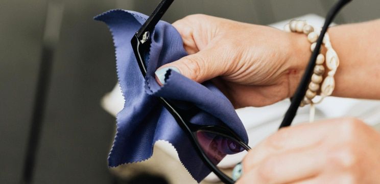A close-up of two hands cleaning a pair of black glasses with a blue cloth. The person has blue nail varnish and is wearing two bracelets on their right hand.