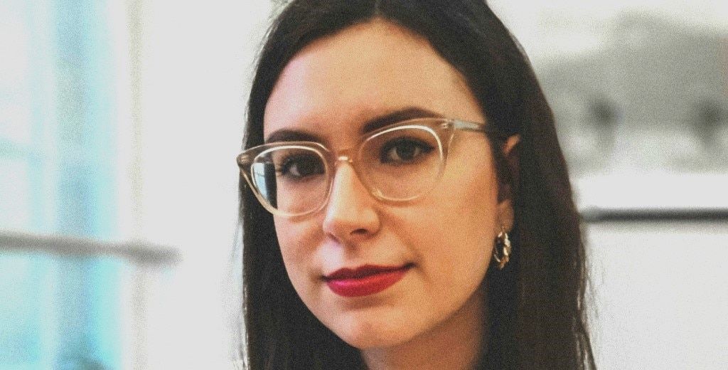 A dark-haired lady in a bright office setting. The background is blurry but you can make out that there is a framed picture and a large window behind her. She is smiling very slightly and has bright red lipstick on. Her glasses are transparent and she is 