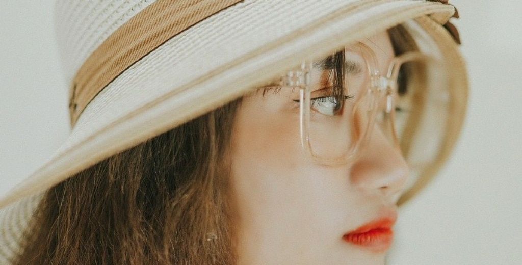 A lady with dark hair wearing oversized clear glasses and a cloche hat. Her lipstick is a bright red colour. She is standing in a brightly lit space and looking to one side, right of the camera.