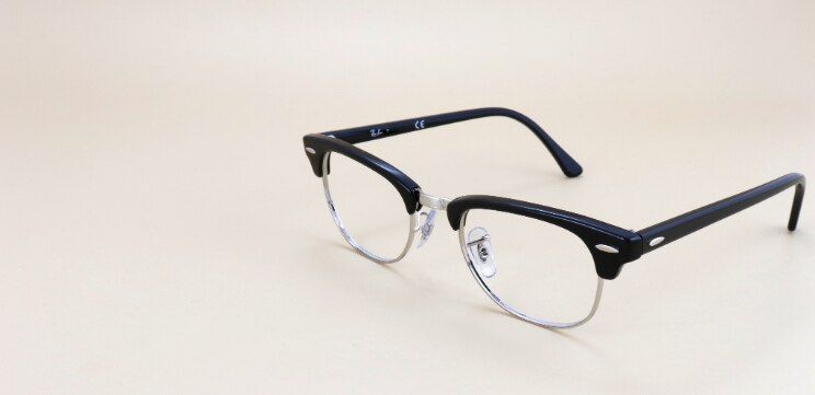 Ray-Ban® Clubmaster glasses in black with silver metal lower rims and pin accents, displayed against a warm neutral background.