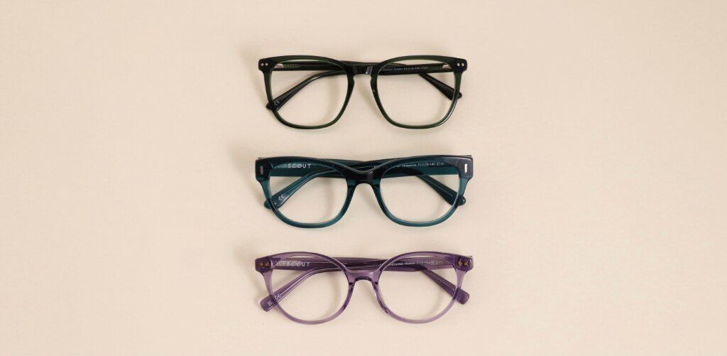 Three Glasses Direct and Scout acetate glasses frames laid flat against a warm neutral background — dark green square, teal cat-eye and transparent purple round, from top to bottom. Frames: GD Elsie, Scout Gracie and Scout Gabriella.