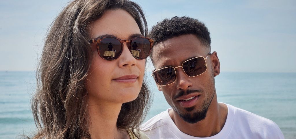 A young couple stands close together on a sunlight beach with the sea in the background. The man, with a short curly undercut, looks at the camera with a slight smile. He’s dressed in a white t-shirt and wears rectangular, gold metal sunglasses. Standing