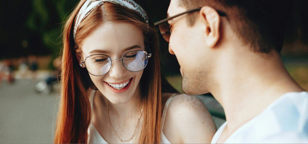 A photo taken over the shoulder of a man as a young couple, both wearing glasses, sit close together outdoors, smiling and laughing, with soft sunlight and a blurred park path in the background. The woman has long straight red hair and is wearing a bandan