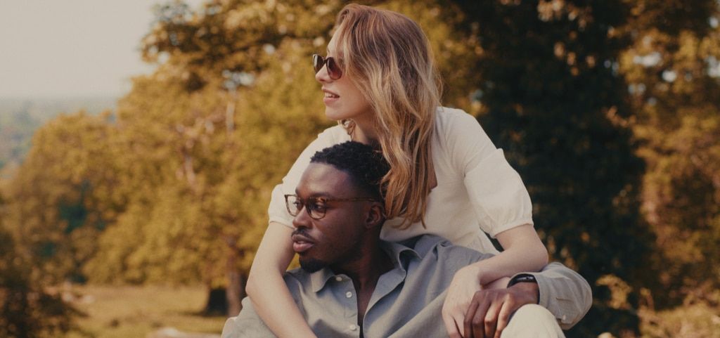 A couple wearing glasses, with the woman leaning forward over the man’s shoulders as they look into the distance, surrounded by sunlit trees in a park-like setting. The woman has long wavy fair hair and is wearing sunglasses and a white mid-sleeve dress, 