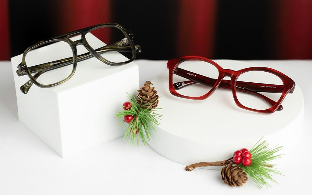 Two pairs of glasses displayed on a smooth white surface against a red curtain backdrop with green fern decorations featuring red berries and pine cones. One pair is a green acetate pilot frame resting on a white cube, and the other is a red square frame 