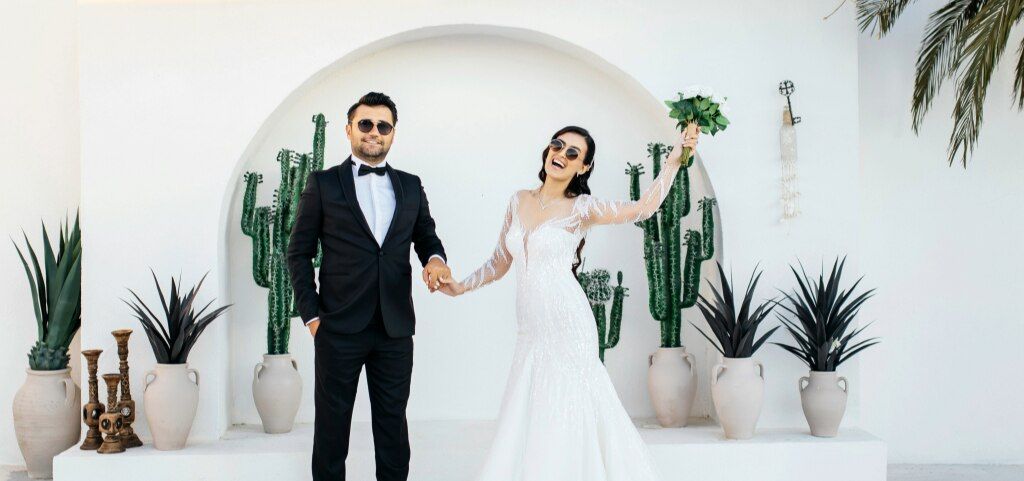 Newlyweds holding hands, standing in a bright white setting with tropical plants, both wearing sunglasses; the groom in a black tuxedo and the bride in a lace fishtail gown holding her bouquet up and smiling.