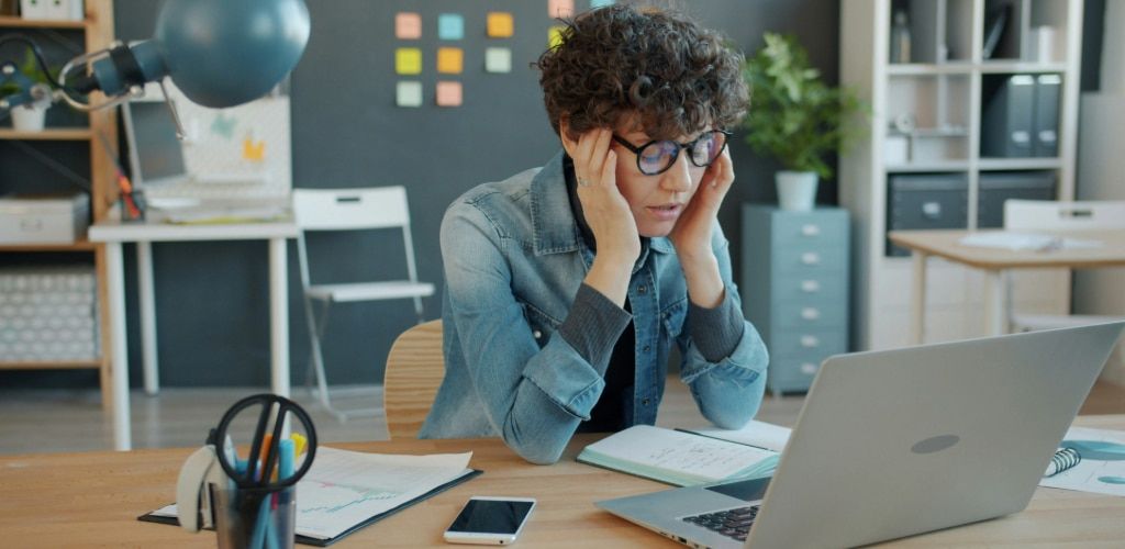 Woman with short curly brown hair wearing round black acetate glasses and a denim shirt over a grey long-sleeve top. She appears to be experiencing digital eye strain while using a laptop, holding her temples and leaning on her desk in a bright, modern of