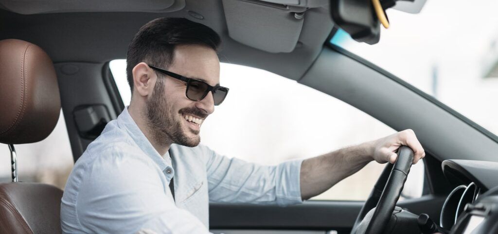 A man wearing sunglasses and white shirt. He is sat smiling in a car with his hand on the steering wheel.
