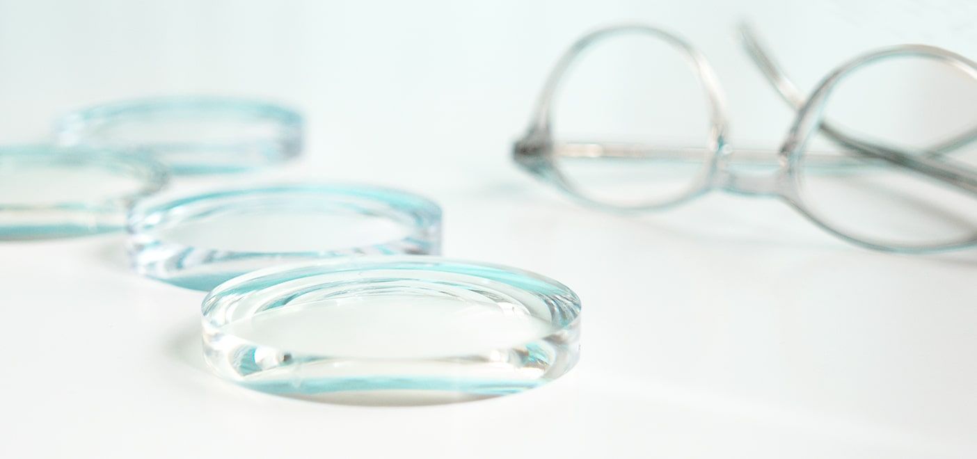 Close-up of thick eyeglass lenses placed on a white surface with a pair of round transparent glasses blurred in the background.