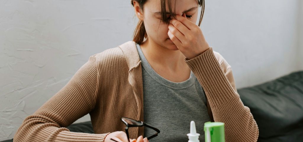 A close-up of a young woman with her hair tied back in a loose ponytail, sitting on a black sofa in an indoor setting. The lady is wearing a grey top and light-brown cardigan. She is rubbing her eyes with her left hand and holding a pair of black glasses 