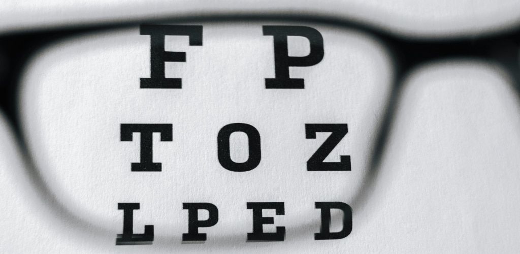 A close-up view of an eye chart seen through the lens of a pair of glasses. The letters inside the lens appear sharp and in focus, while the surrounding areas are blurred, illustrating how corrective lenses improve vision.