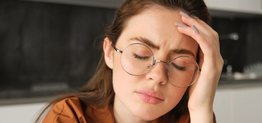 A close shot of a young lady holding a hand to her head with her eyes closed. Her brown hair is partly swept back and she is wearing a pair of large silver, metal round glasses and a dark-orange top.