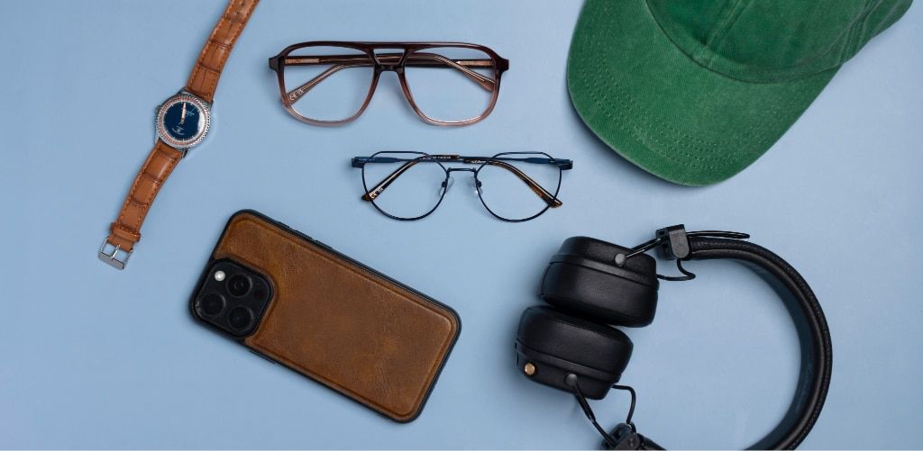 Flat lay of two pairs of glasses, including brown gradient plastic aviator frames and blue metal phantos glasses, alongside a watch with brown straps, smartphone in a brown leather-style case, over-ear headphones and a green baseball cap, illustrating an