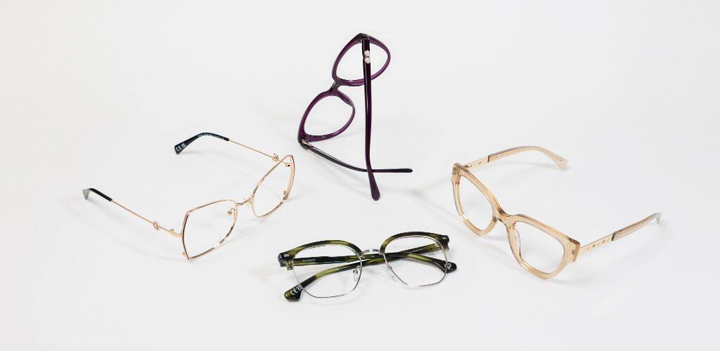 Four pairs of glasses arranged on a white surface, including Scout Kenna gold butterfly metal frames, Scout Kyro purple cat-eye frames, Scout Keegan green square browline frames and Scout Kenzie transparent beige squared cat-eye frames with embellished me