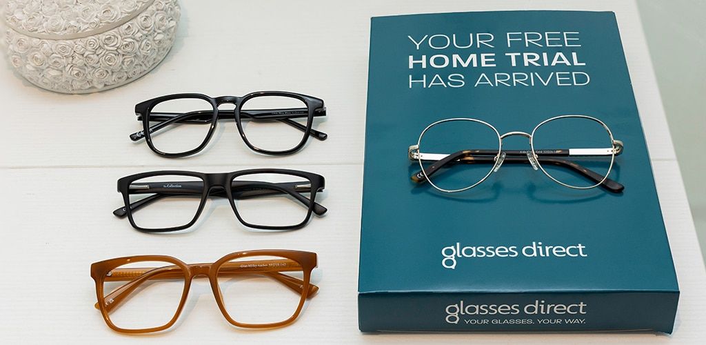 Three pairs of glasses beside a Glasses Direct Home Trial box on a white table, including black rectangular frames, square black frames, orange square frames, and round gold metal glasses resting on top of the box.