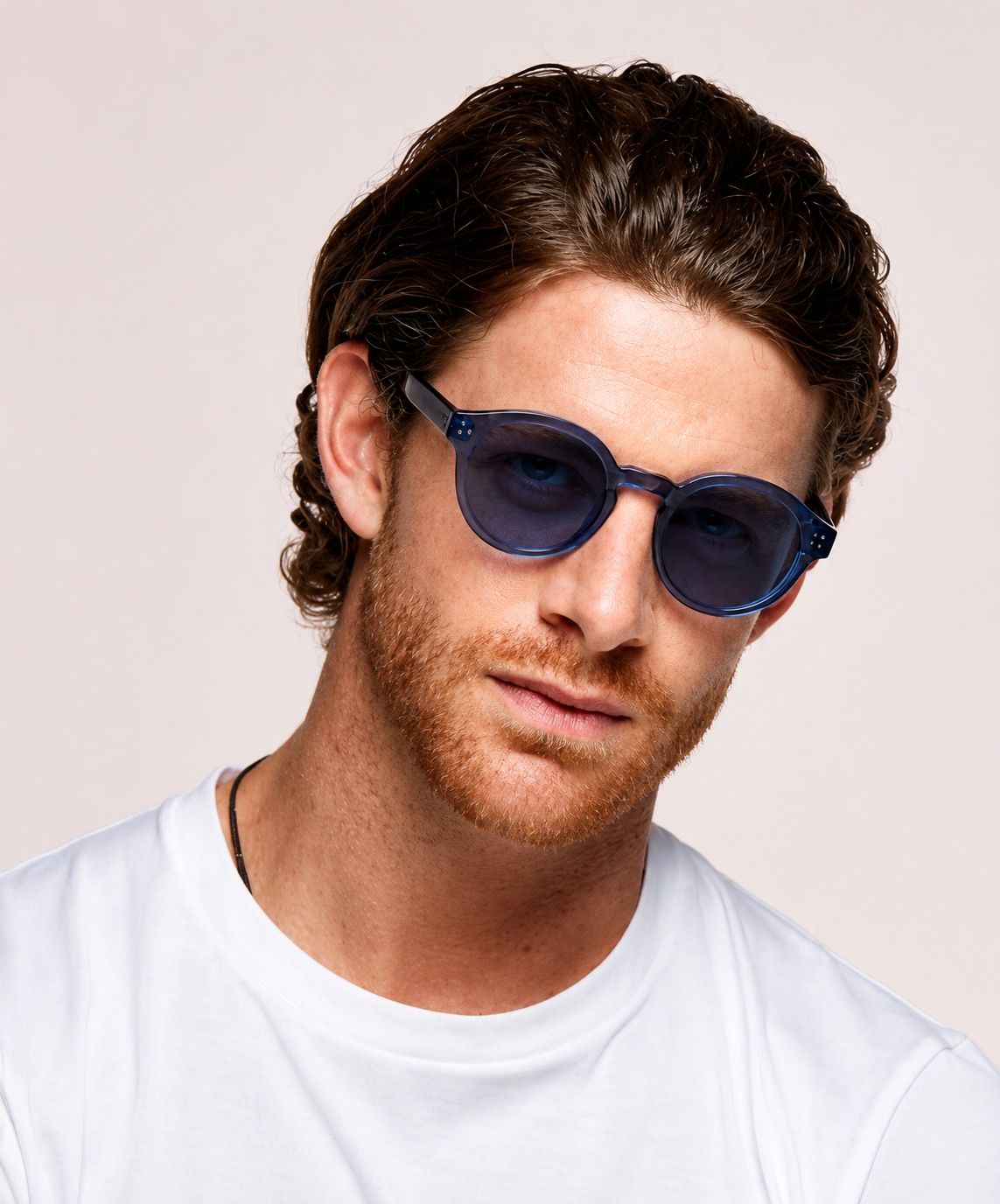 A man with medium-length wavy brown hair and light facial hair wears large round blue sunglasses with amethyst-tinted lenses and a white t-shirt. He tilts his head slightly and looks toward the camera with a relaxed expression against a plain light backgr