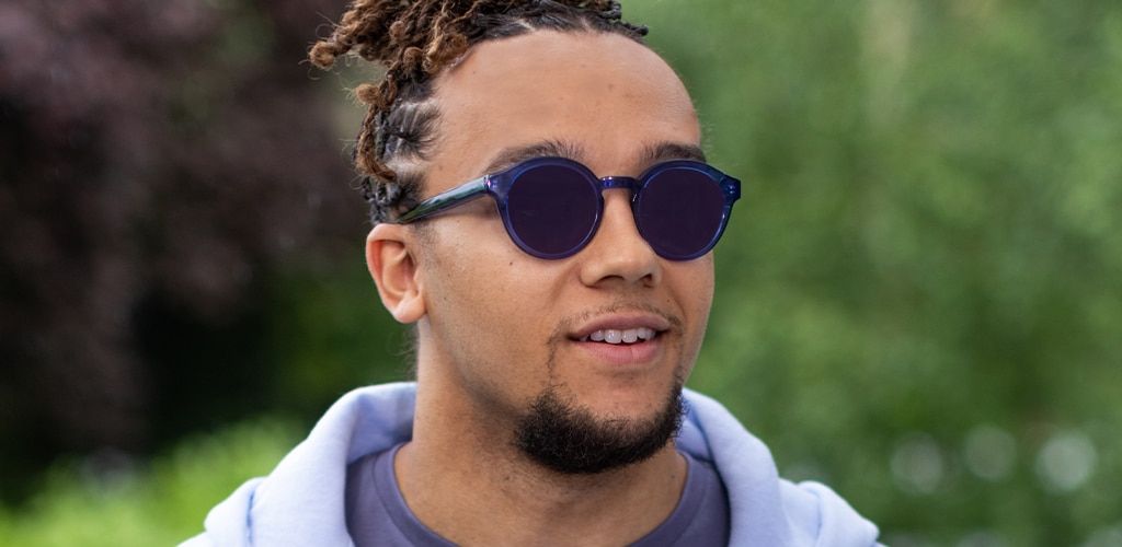 Close-up of Perri Kiely smiling outdoors, hair tied in a bun and sporting a goatee, wearing bright-blue round sunglasses with Transitions® GEN S™ amethyst lenses, a light blue t-shirt, and a baby blue hoodie.