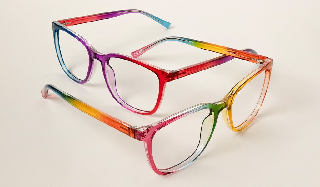 Two rainbow-coloured square acetate glasses lie intertwined on a cream backdrop, each pair open and angled slightly apart.