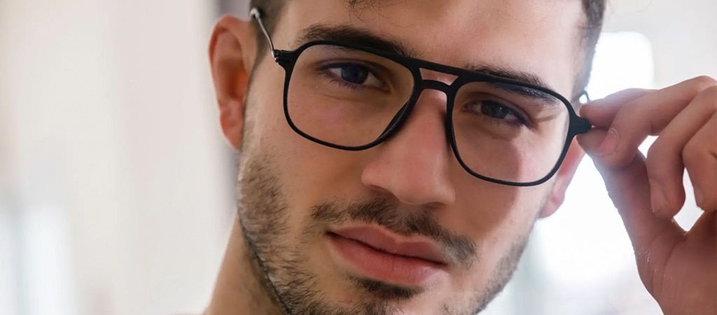 A close-up of a young man with dark hair and light stubble wearing black pilot-style geometric glasses. He is in a bright indoor setting. His head is tilted to one side and he is holding the edge of his glasses with his left hand.