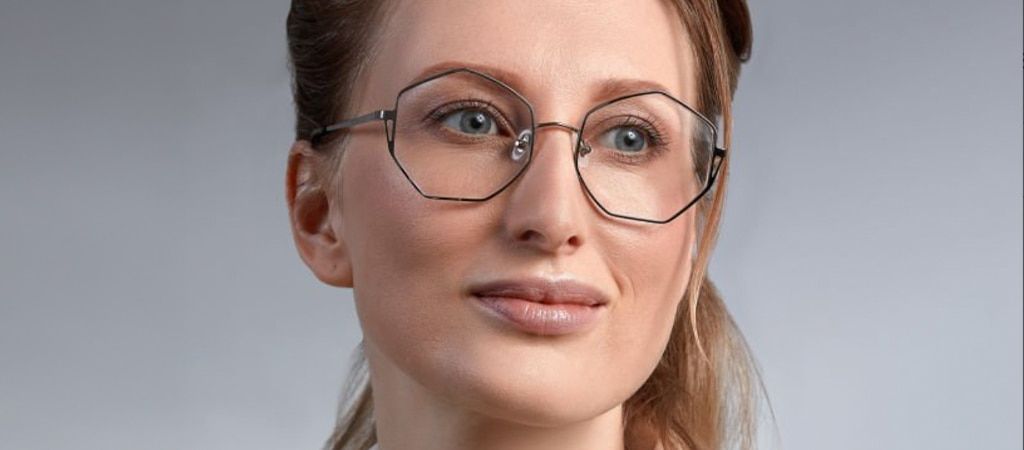 A close-up of a lady with dark-blonde hair tied back off her face. She is wearing a pair of black metal geometric glasses and looking to one side. The background is white and shadowy.