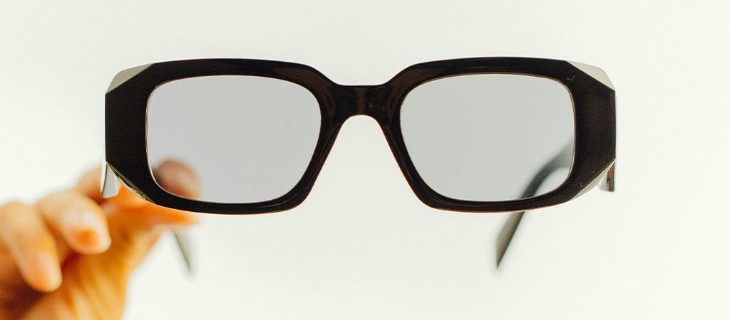 A pair of black acetate hexagonal glasses held up on a cream background. The hand holding them can be seen out of focus to one side.