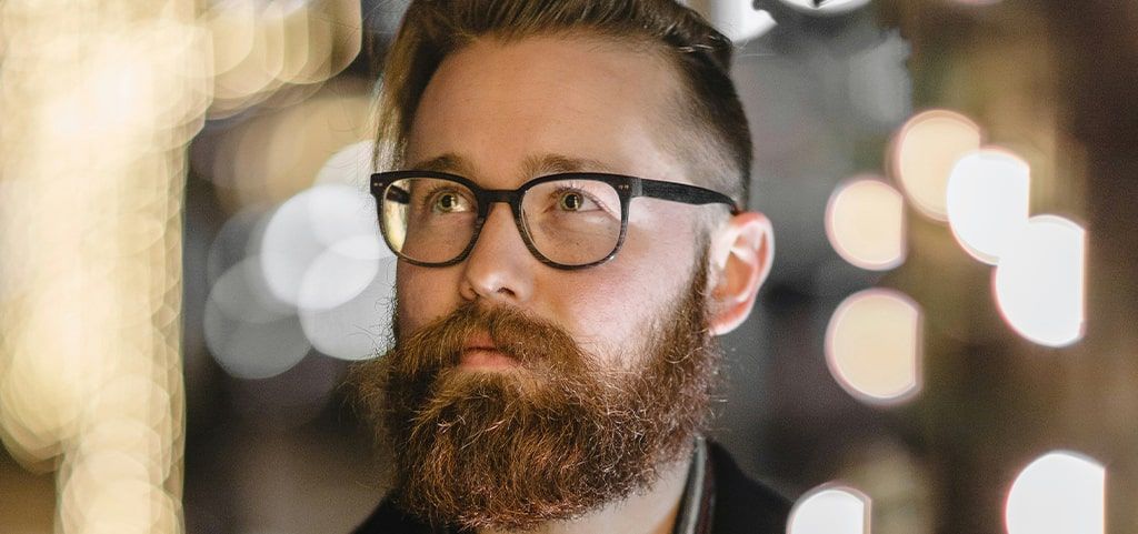 A close up of a man with a full beard wearing a pair of black acetate round glasses as he looks off to the side with a calm expression, shown in a close-up portrait with soft lighting and a neutral background.