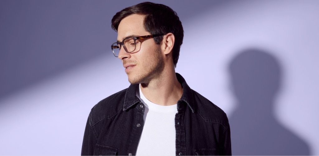 A man with short dark hair wearing tortoiseshell rectangular glasses stands against a soft lavender background with dramatic studio lighting. He's dressed in a dark denim-style jacket over a white T-shirt and is looking slightly downward and to the side. 