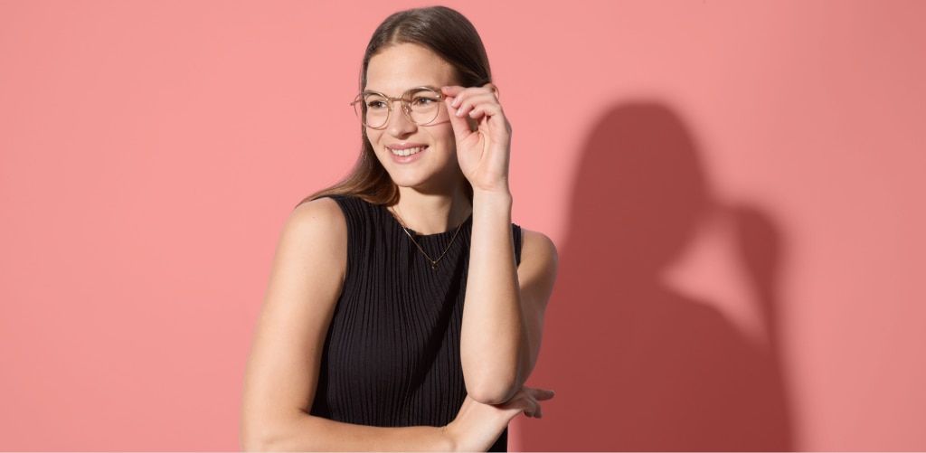 A smiling woman with long brown hair wearing thin metal round glasses poses against a soft pink background. She's dressed in a sleeveless black ribbed top and lightly touches the corner of her glasses with one hand while looking off to the side. Strong st